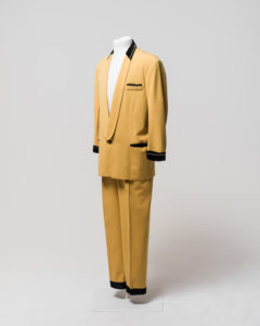 Men's suit consisting of jacket and trousers, worn by Johnny O'Keefe. -1 Jacket made of yellow [cotton] fabric with black velvet trimming at collar, cuffs and pockets. The jacket has a shawl collar with the neck area trimmed with black velvet decorated with a string of diamantes around the neck. The lapels are of the same yellow fabric and extend to the single button fastening at the waist. The button is made of brownish-black plastic. The jacket is long sleeved and the cuffs are trimmed with black velvet decorated with a string of diamantes around the front half of both cuffs, some diamantes are missing from both cuffs. The jacket has a fake external left breast pocket trimmed with black velvet and diamantes, one diamante appears to be missing from the end of the string. The jacket also has two fake external waist pockets on the left and right sides which are trimmed with black velvet. The jacket is lined with salmon pink silk and has no internal pockets, the shoulders have some padding. A maker's label is sewn inside the jacket at the left breast, the label is cream coloured with black embroidered text "Len Taylor/Sydney/American Clothes Stylist". The label is partly detached from the jacket. The jacket has black marks exteding horizontally from just above the right waist pocket and discolouration in the lining at the centre back neck area and in various places on the external yellow fabric. There are several moth holes in the lining and in the external fabric particularly just below the right waist pocket. -2 Trousers made of yellow [cotton] fabric, matching trousers to jacket (98/32/1-1). Trousers have 2 side pockets and are pleated at the front of waist. Trousers fasten at centre front with a metal zipper, metal hook and eye and internal button which fastens to button hole on a triangular extension of yellow fabric (this internal button is missing). There are seven belt loops around waist for a thin belt. Trousers are unlined except for the inside waist which is lined with a band of ribbed cream silk, covering a rougher piece of hessian coloured fabric, the zipper is also lined with cream silk. The waistband lining is partially detached from the trousers. The legs of the trousers are hemmed with black velvet giving the appearence of a cuff at the hem of each leg. The cream cotton fabric forming the inside of the left pocket has a handwritten inscription in black ink "J.R O'Keef" [sic] and the fabric forming the inside right pocket has a handwritten inscription in black ink "FHC".